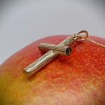 A solid 9 karat gold cross pendant with a distinctive 3D log-on-log structure, hand-engraved with a rich tree bark texture. Leaning right and resting on a mango, the warm fruit tones highlight the pendant’s natural detail and layered form—blending organic elegance with lasting craftsmanship.