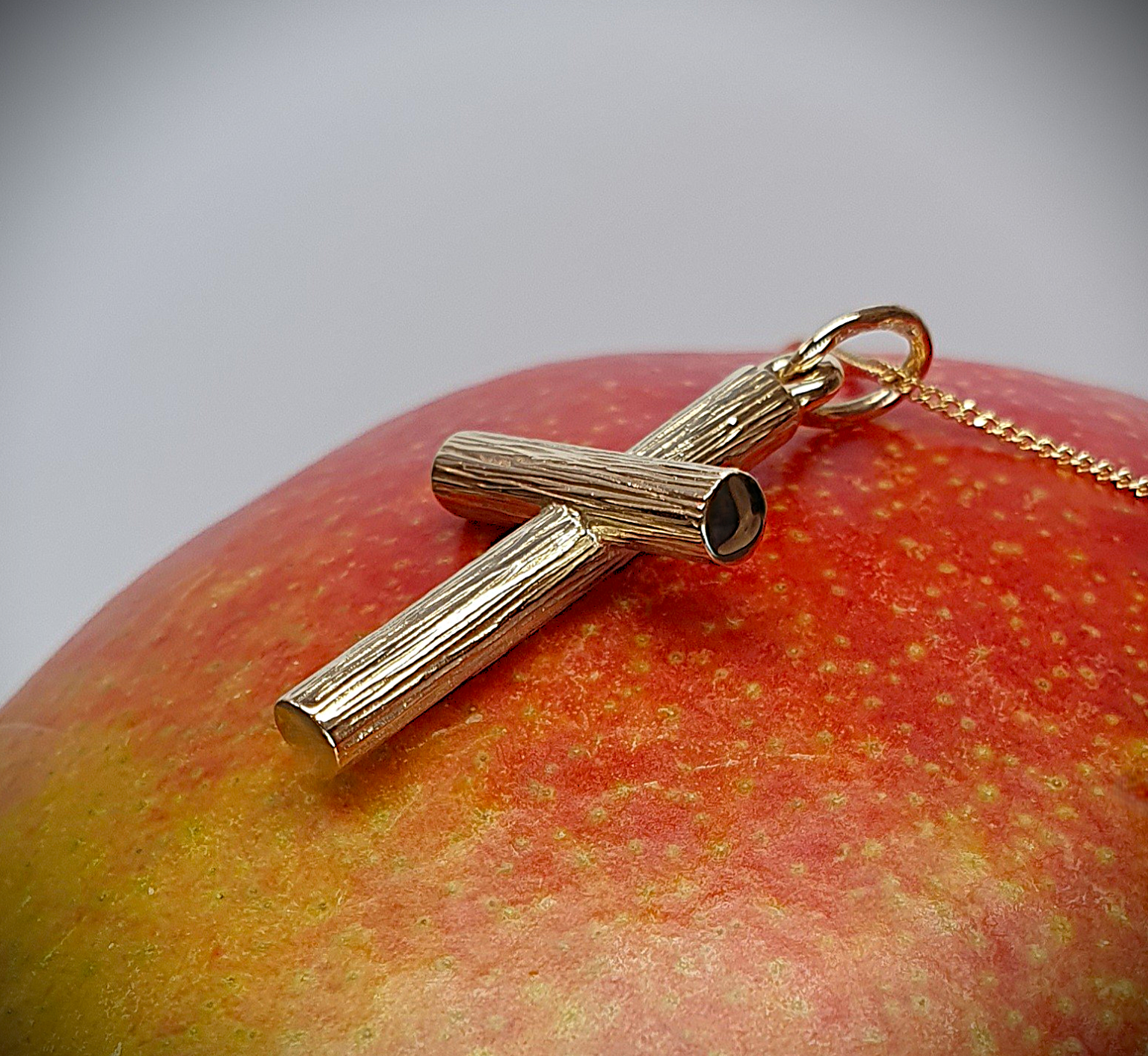 A solid 9 karat gold cross pendant with a distinctive 3D log-on-log structure, hand-engraved with a rich tree bark texture. Leaning right and resting on a mango, the warm fruit tones highlight the pendant’s natural detail and layered form—blending organic elegance with lasting craftsmanship.
