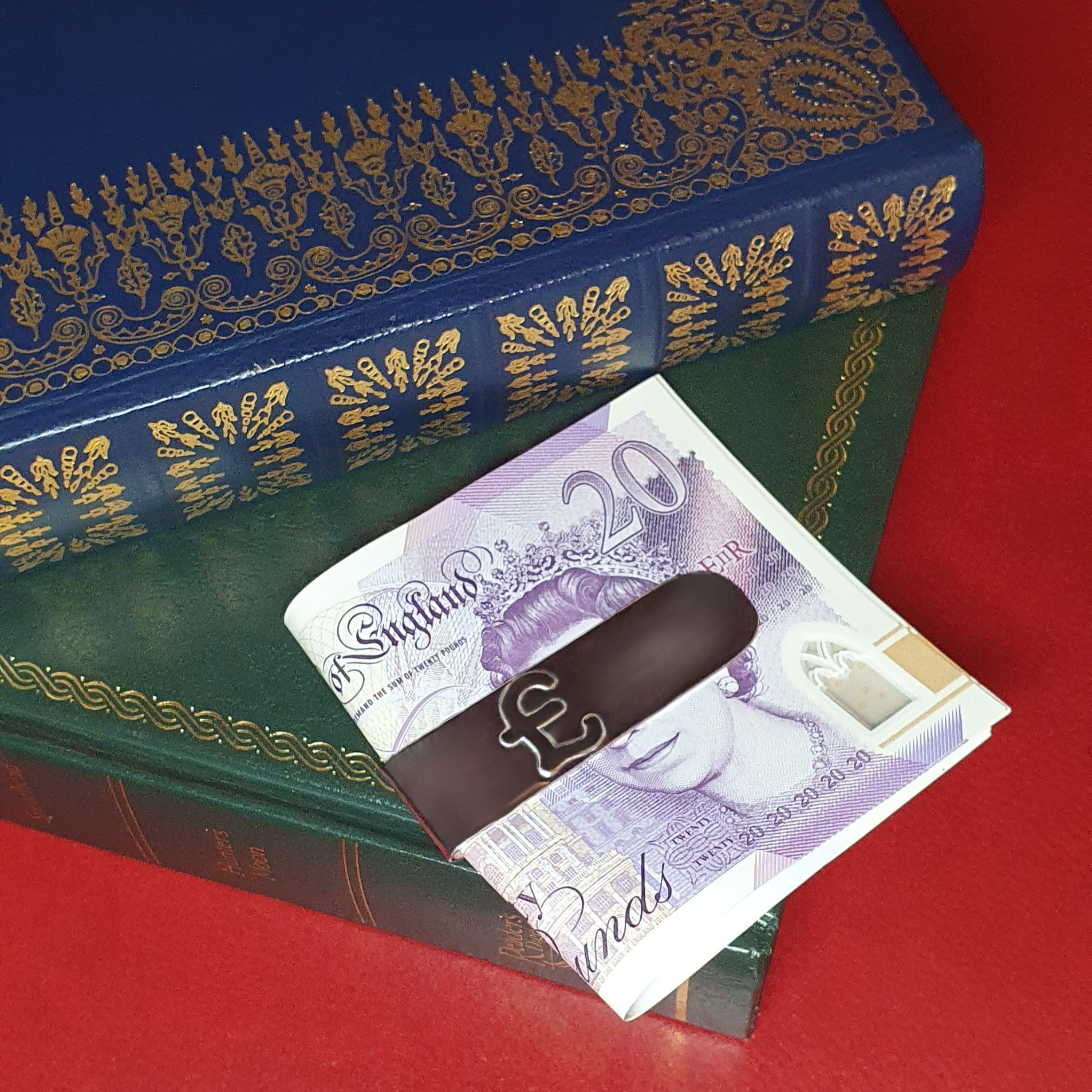The front of the real silver Pound sign money clip is pictured with a twenty pound note on a green and blue leather book.