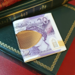 A solid gold cash clip is pictured on twenty-pound notes on top of some books.