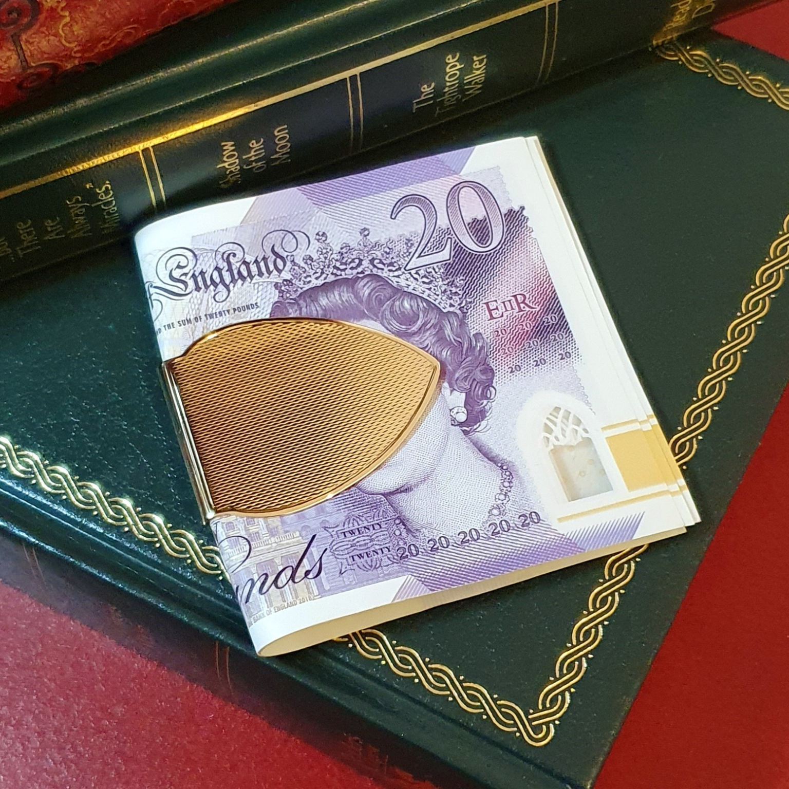 A solid gold cash clip is pictured on twenty-pound notes on top of some books.