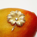 A textured yellow gold leaf brooch pin set with a single sparkling white diamond in the centre, pictured on a mango.