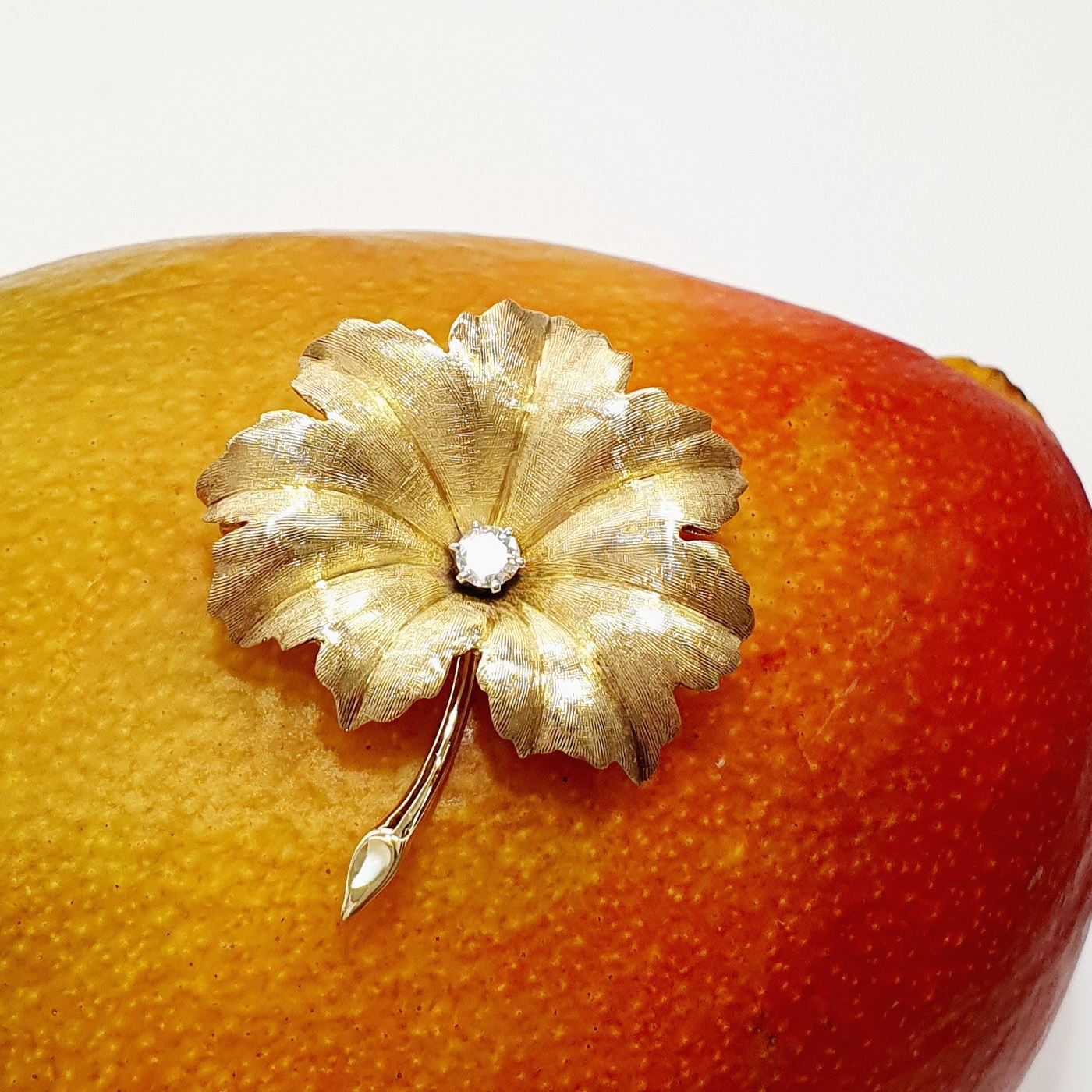 A textured yellow gold leaf brooch pin set with a single sparkling white diamond in the centre, pictured on a mango.