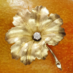 A textured yellow gold leaf brooch pin set with a single sparkling white diamond in the centre, pictured close up on a mango.