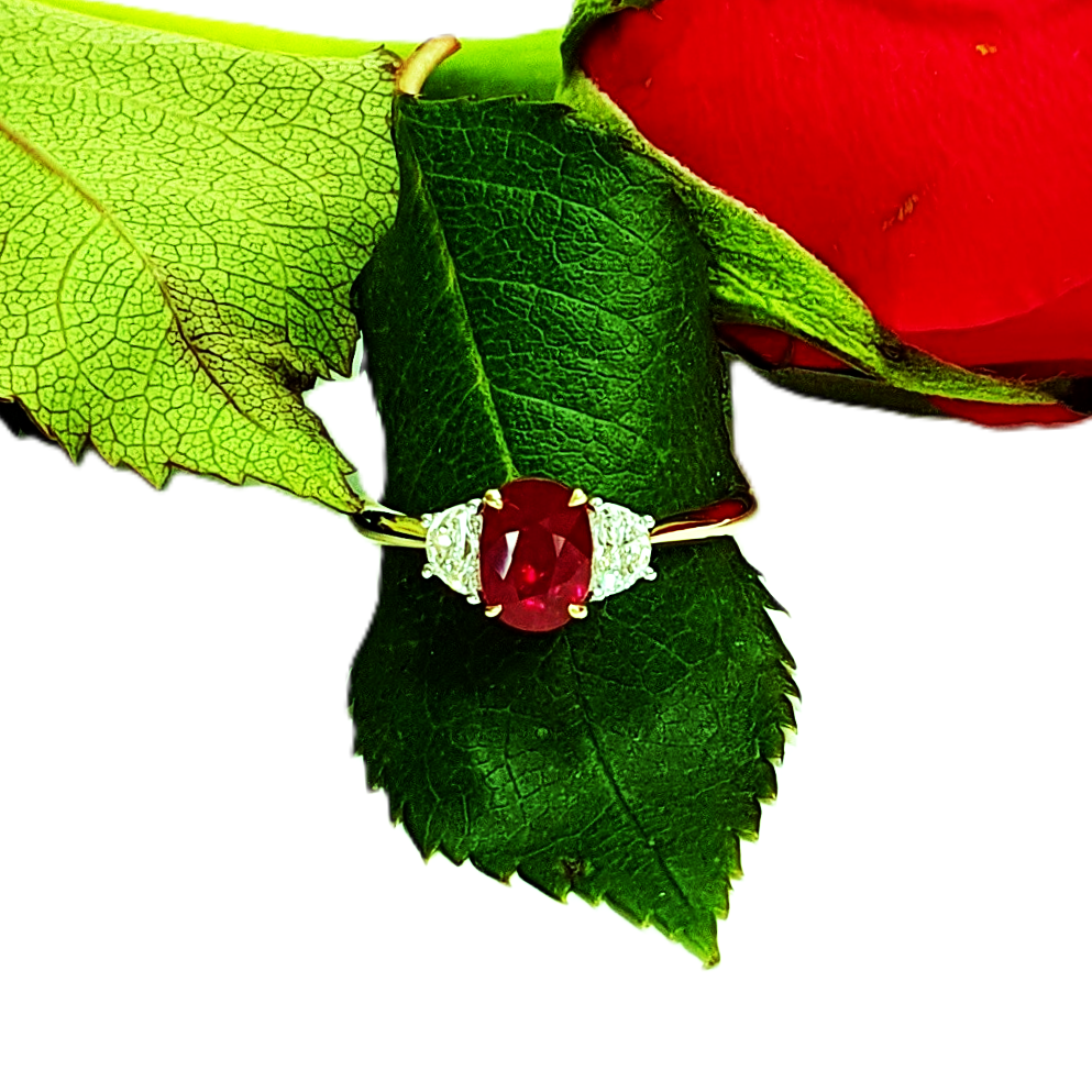 18ct yellow gold three-stone ring with one oval ruby and two half-moon diamonds set in 18ct white gold, one on each side, pictured on a single red rose leaf RA00698.