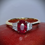 An 18ct yellow gold oval ruby and two half-moon diamond ring with white gold settings on the diamonds is pictured close up on a red mango, RA00698.