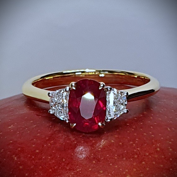 An 18ct yellow gold oval ruby and two half-moon diamond ring with white gold settings on the diamonds is pictured close up on a red mango, RA00698.