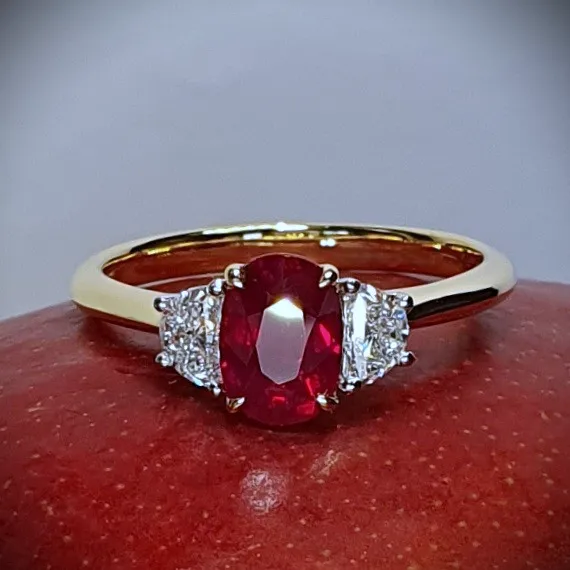 An 18ct yellow gold oval ruby and two half-moon diamond ring with white gold settings on the diamonds is pictured close up on a red mango, RA00698.