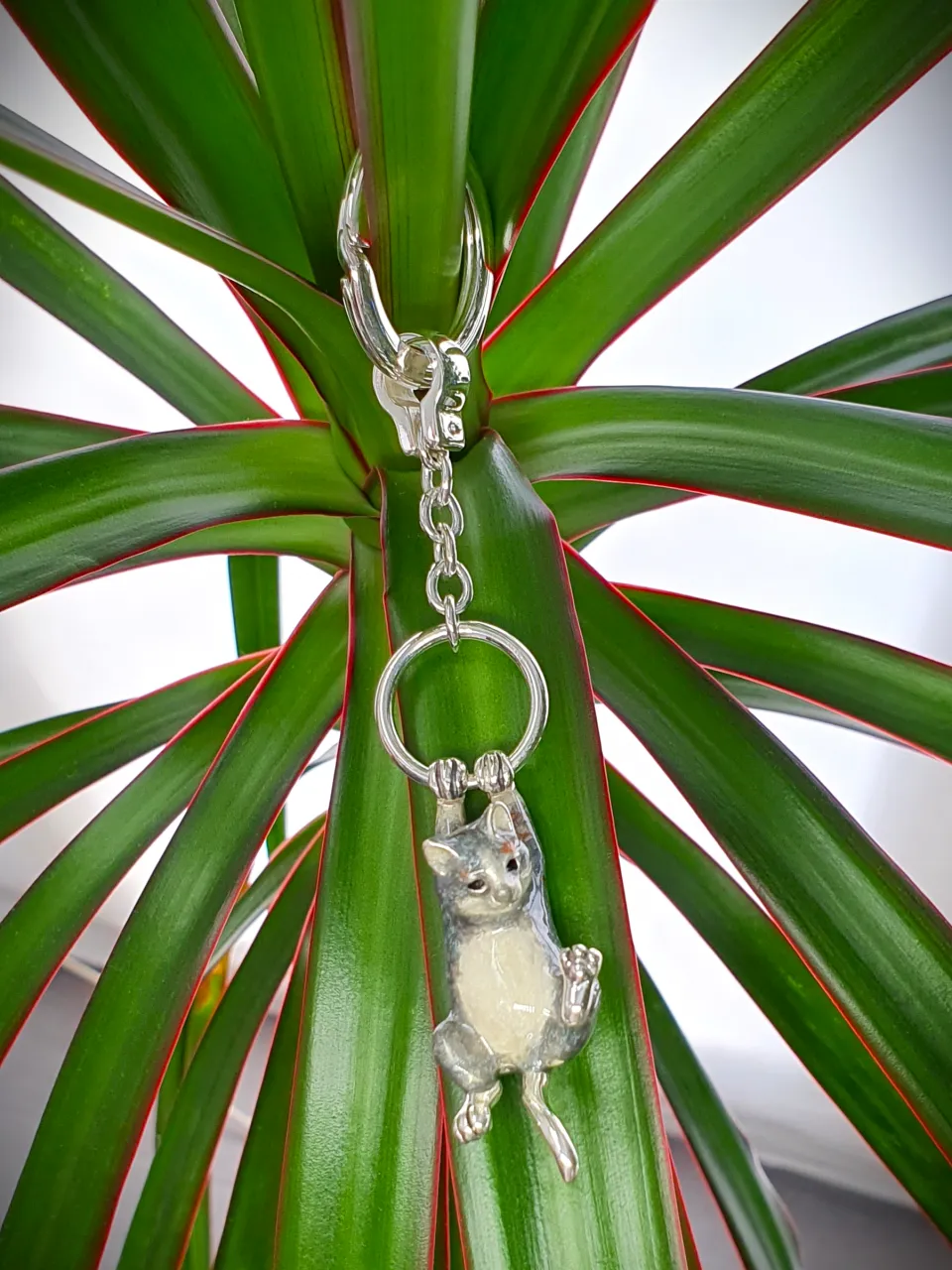 Genuine hallmarked silver tabby cat keyring fob or handbag charm with a grey brown life-like enamelled finish KF00048.Pictuired hanging from a plant.