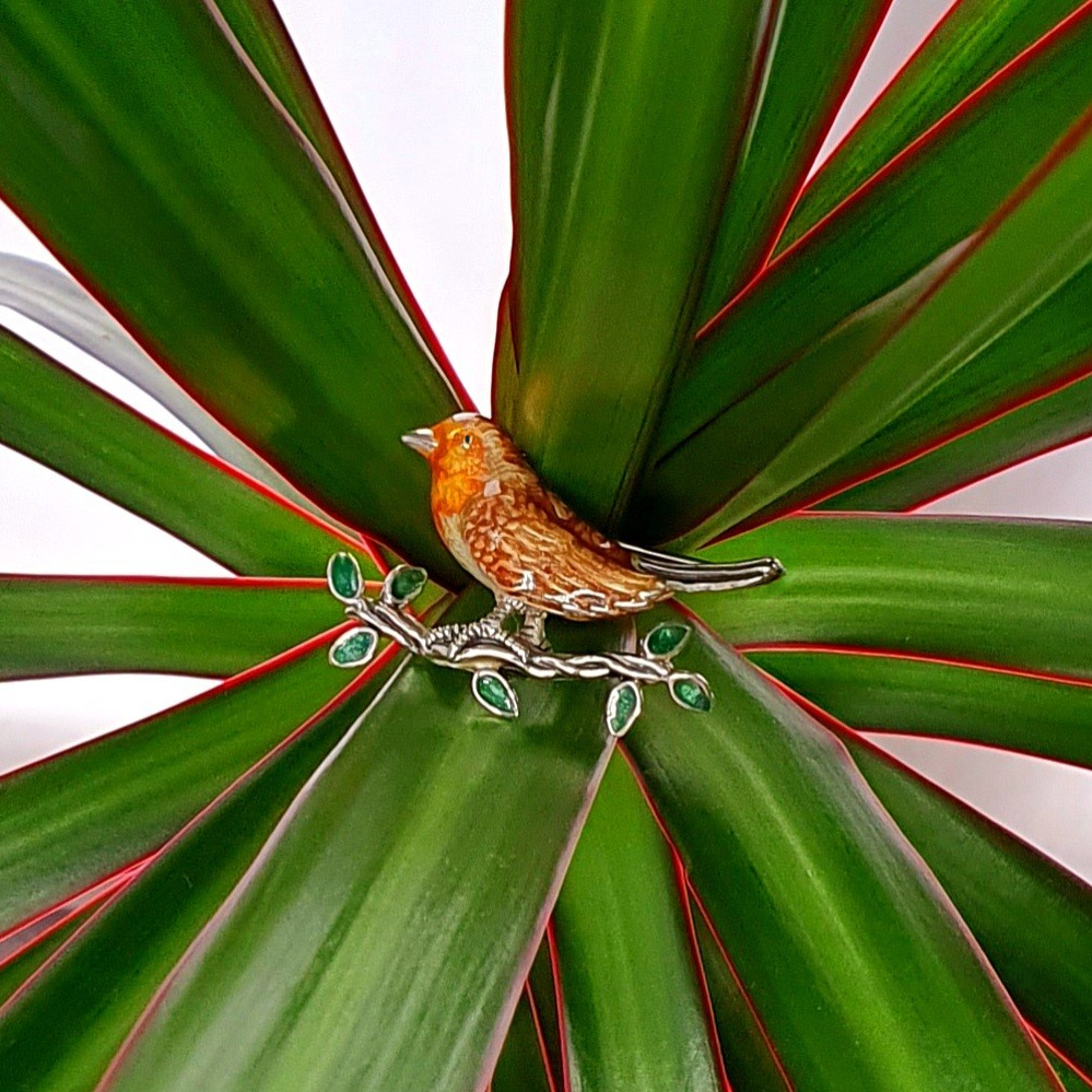 Saturno sterling silver enamelled robin red breast brooch sitting on a branch with enamelled green leaves pictured on a plant.