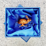 In-box: Running hare brooch nestled in a blue silk-lined gift box, its dynamic pose and enamelled finish gleaming under soft folds of the silk.