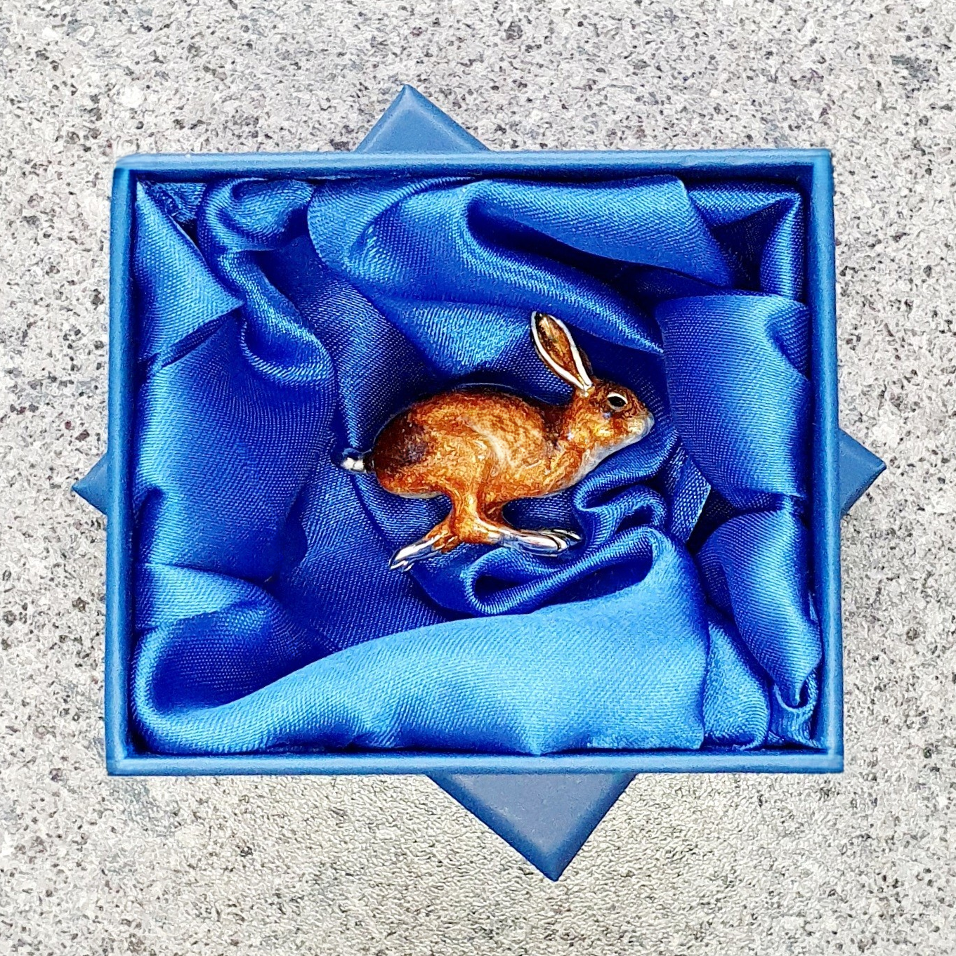 In-box: Running hare brooch nestled in a blue silk-lined gift box, its dynamic pose and enamelled finish gleaming under soft folds of the silk.