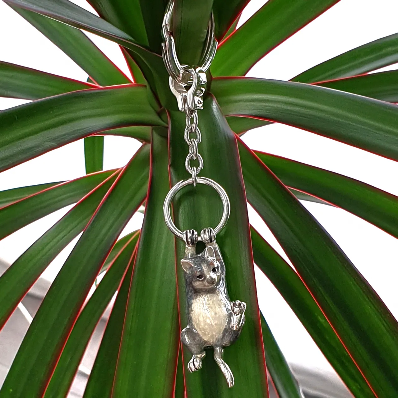 Genuine hallmarked silver tabby cat keyring fob or handbag charm with a grey brown life-like enamelled finish KF00048.Pictuired hanging from a plant.
