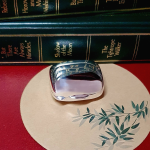Closed pill box beside stacked books on bamboo-themed placemat Shimmering silver finish beautifully reflects natural light while seated on a round beige mat with green bamboo illustrations.