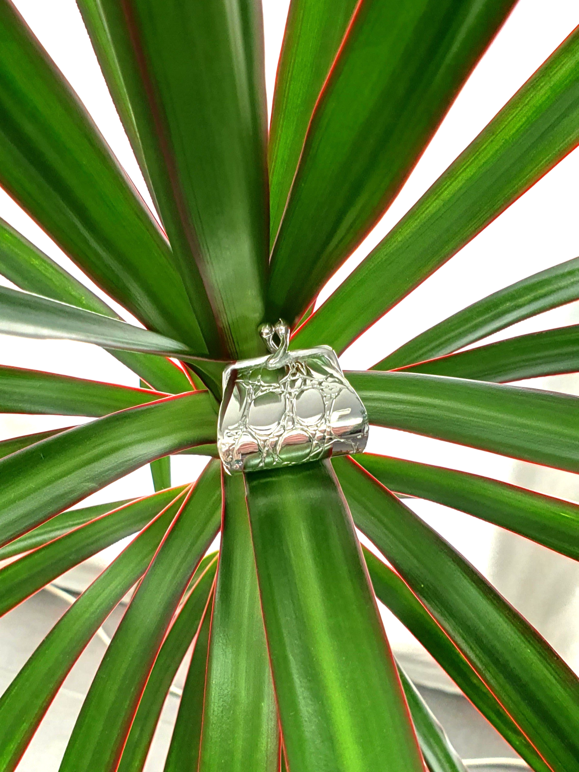 Sterling silver handbag pill box balanced on green leafy plant, blending organic texture with luxury miniature design.
