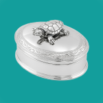 Oval silver box with scroll-engraved lid and movable turtle, set against a turquoise backdrop.