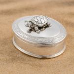 Silver oval pill box with scroll engraving and movable turtle, photographed on soft sand.
