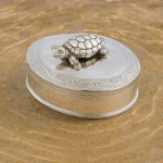 Sterling silver box with scrollwork and movable turtle, placed on sand beside rippling water.