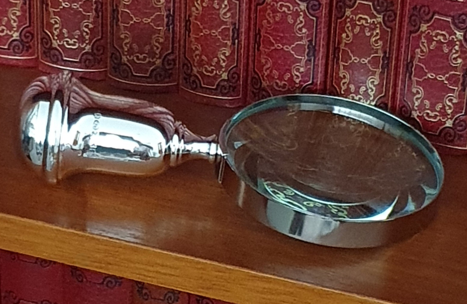 Close-up of sterling silver magnifying glass handle, angled on bookshelf with red leather-bound books in background.8