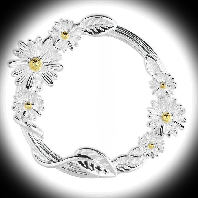 A dramatic black‑background image of the Sterling Silver Daisy Circle Brooch illuminated by a soft white spotlight at the centre. The two‑tone daisies glow warmly, while the wrapped stem and polished silver circle create a striking contrast.