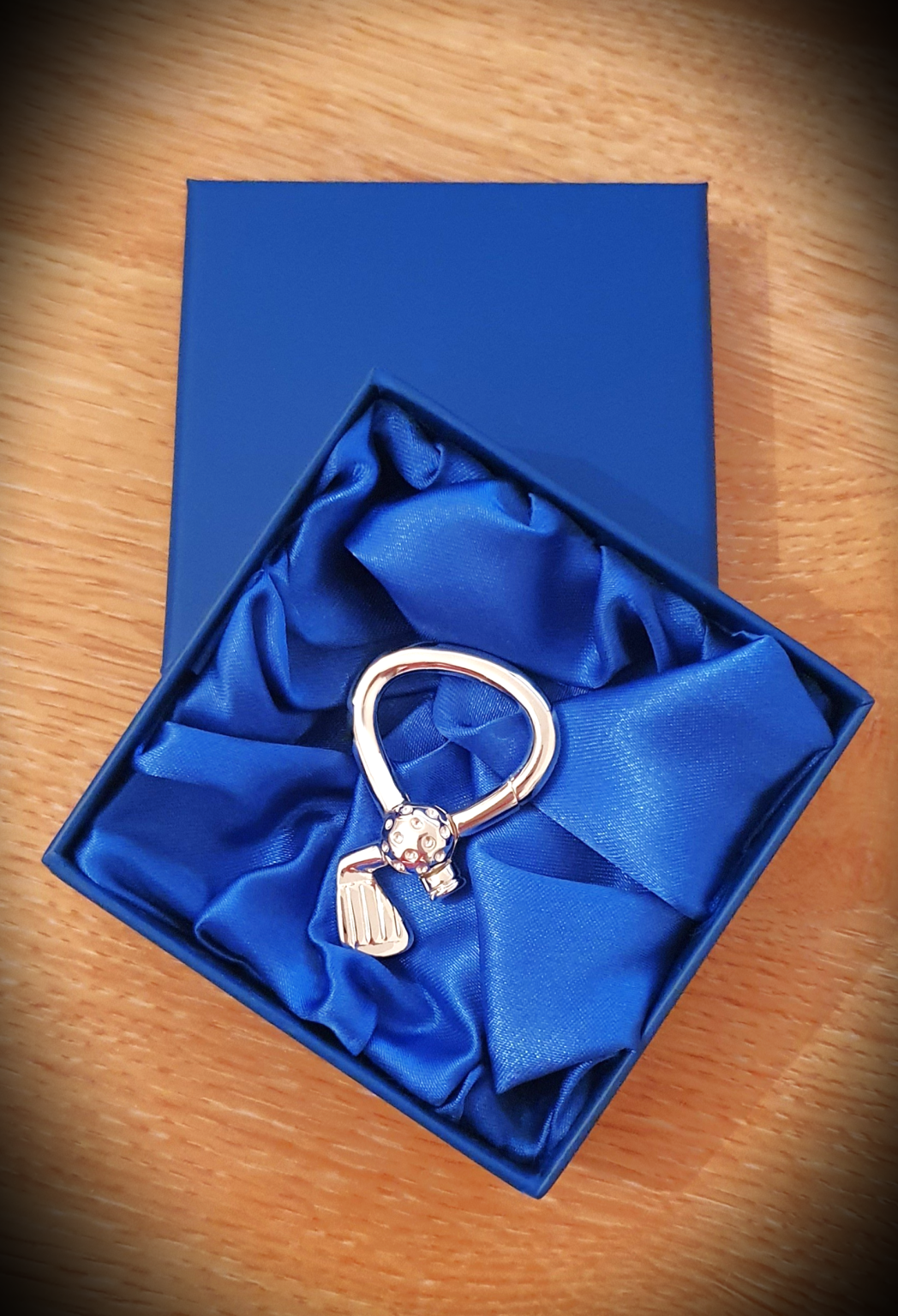 The real silver golf club and ball key ring rests in a deep blue presentation box lined with soft blue silk. The left‑facing angle and rich fabric texture enhance the shine of the club and the detailed ball.