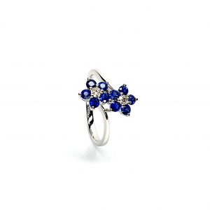"An elegant 18ct white gold ring with a crossover shank featuring two clusters of five round bright blue sapphires, each cluster surrounds a single white sparkling diamond, creating a captivating and sophisticated design." Angled View.