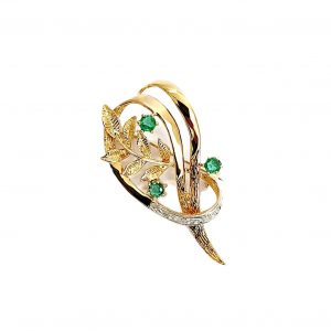 9ct yellow gold brooch with a sprig of seven leaves coming through a floral swirl of polished and barked yellow gold with three bright green emeralds & three sparkling diamonds set into a small white rhodium section, top view.